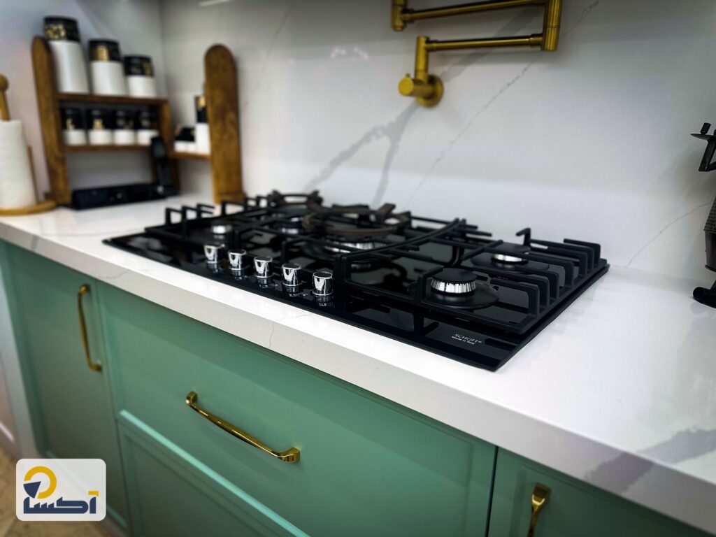 اجاق گاز مشکی روی صفحه کوارتز سفید و کابینت‌های زمینی سبز با دستگیره طلایی